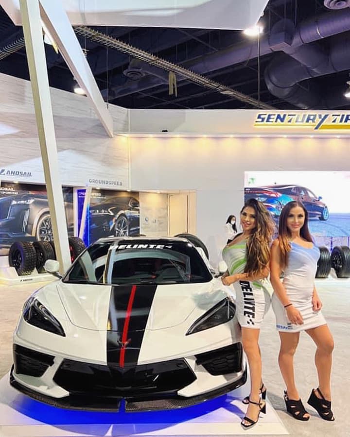 Two brand ambassadors in branded Delinte dresses pose beside a white Corvette at a Delinte/Sentury Tire trade show booth.