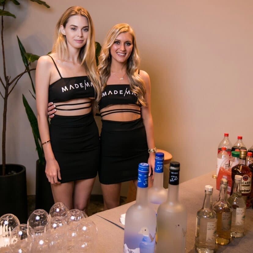 Two brand ambassadors in black 'MADE' crop tops and skirts pose behind a bar stocked with liquor bottles and glassware.