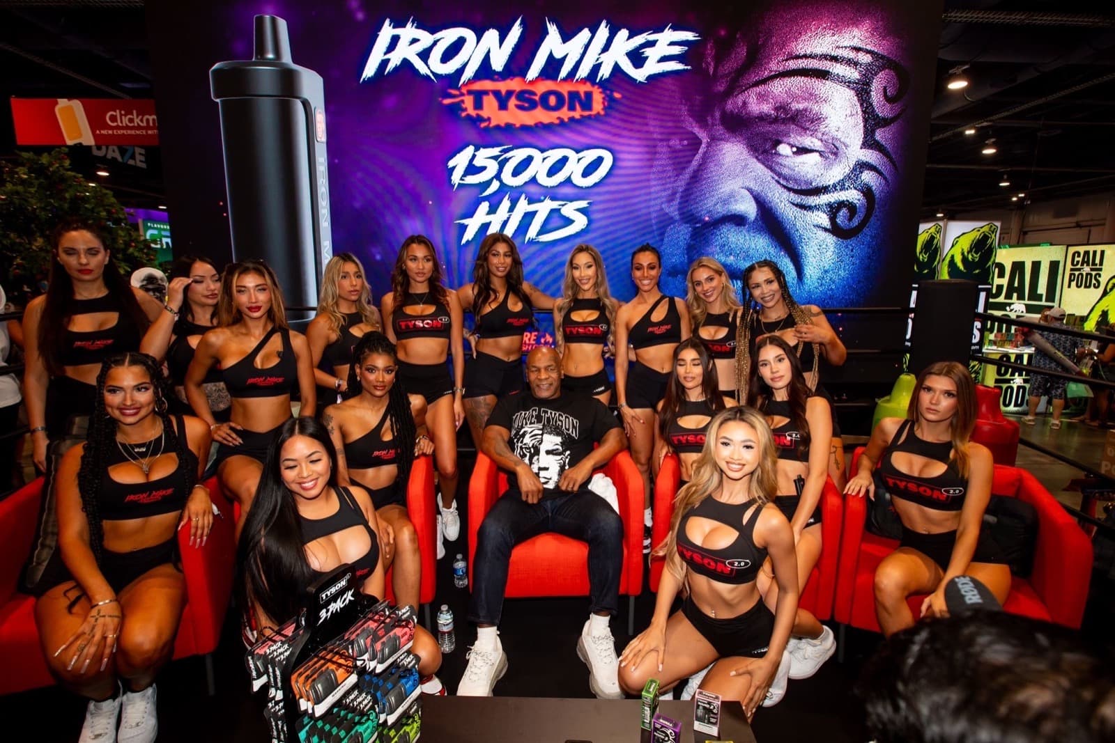 More than a dozen brand ambassadors in matching black 'Iron Mike' crop tops and shorts surround a seated Mike Tyson at an Iron Mike Tyson vape trade show booth, with product 3-packs displayed in front and a large screen backdrop.